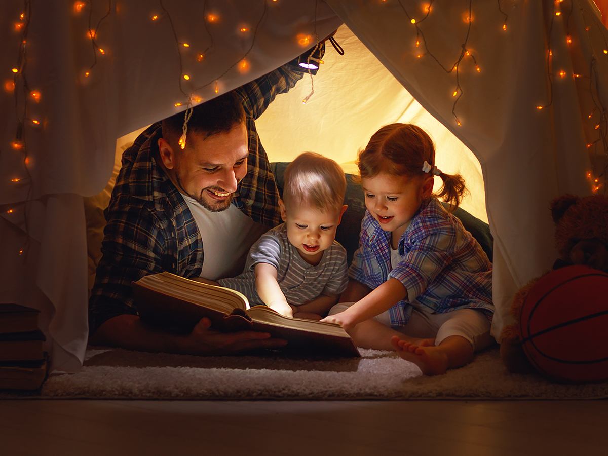 Dad reading in blanket fort with 2 toddler daughters